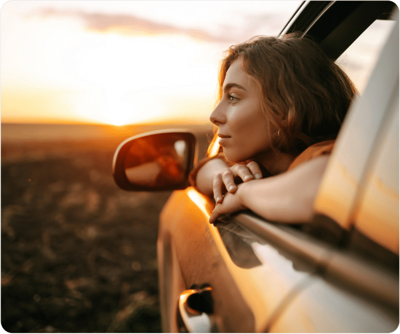 A person leans out of a car window, enjoying a sunset over a vast landscape, with warm light reflecting off the vehicle.
